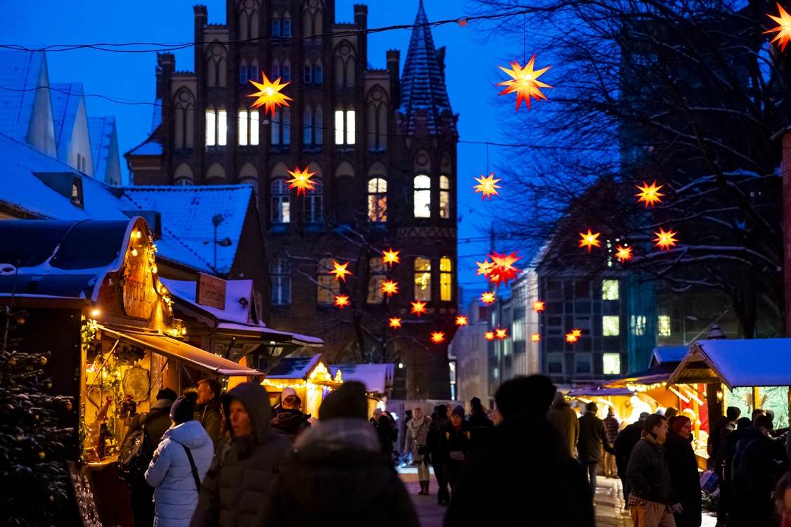 Beleuchtete Stände auf einem Weihnachtsmarkt bei Nacht mit leuchtenden gelb-roten Sternen an Lichterketten über dem Markt und historischen Gebäuden im Hintergrund.