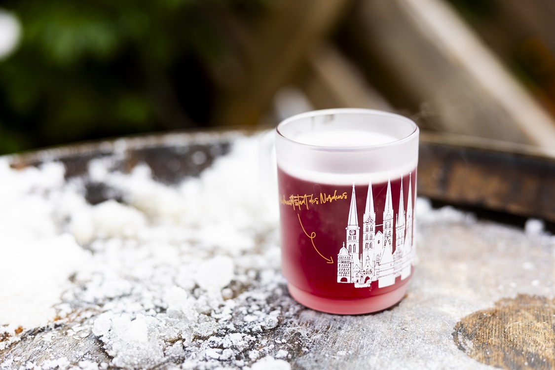 Glühweintasse mit Lübeck-Motiv steht auf einem verschneiten Holzfass.