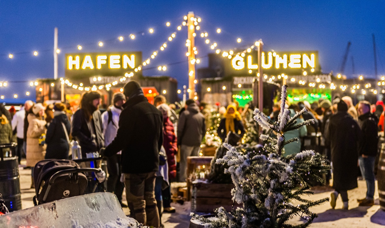 Menschen auf einem Weihnachtsmarkt mit Lichterketten, Tannenbäumen und leuchtendem Schriftzug „Hafen Glühen“ über zwei Buden.